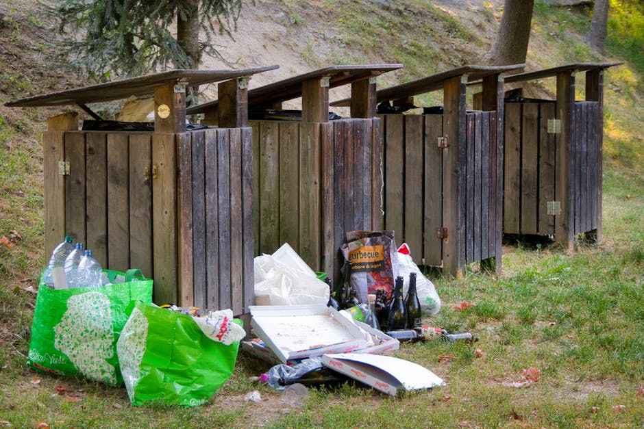 Four wooden waste bins with slanted roofs, arranged in a row on a grassy area, are used for rubbish storage and disposal. The bins are made of vertical wooden planks, stained dark brown, with metal hinges and handles visible on the doors. In front of the bins, there are various discarded items, including a green plastic bag filled with empty plastic bottles, a white cardboard pizza box with some residual food, a black plastic bag, several glass bottles, and other debris scattered on the grass. Surrounding the bins are trees and a hillside covered with grass and soil, creating a natural outdoor environment. The scene suggests a private waste collection area, possibly for a nearby property, with visible signs of recent rubbish disposal. The overall atmosphere indicates an outdoor setting dedicated to waste management or rubbish collection, with House Clearance Belsize Park providing services related to on-site clearance and waste removal in a residential area.