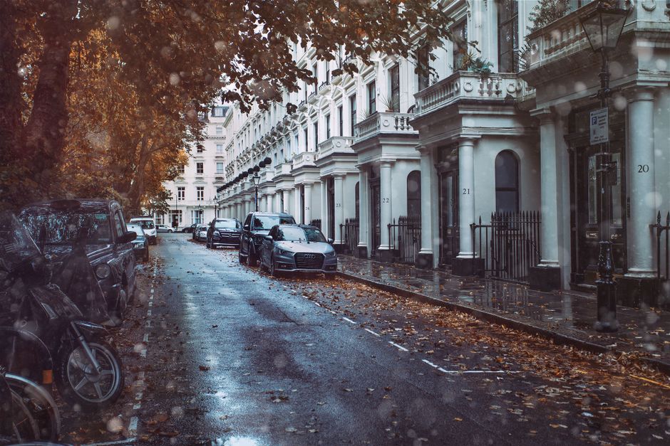 A residential street scene showing a narrow asphalt road with wet patches and scattered fallen leaves, indicating recent rain, and lined with parked cars on both sides. The vehicles include a black van, a dark grey sedan, and a black motorcycle, positioned close to the curb on the left side. On the right side, there are white, ornate terraced townhouses with large arched windows, decorative mouldings, and small front gardens enclosed by black wrought-iron fences, each numbered from 21 to 24. Tall street lamps with traditional lantern-style fixtures are situated along the pavement adjacent to the buildings. Overhanging trees with dense foliage exhibit rich autumnal hues of orange and brown, partially obscuring the upper floors of the buildings. The scene is lit by diffused natural daylight, reflecting off the wet surfaces, and conveying a quiet, urban residential atmosphere. The presence of parked cars and urban infrastructure subtly connects to the context of private waste handling or alternative rubbish collection services offered by companies like House Clearance Belsize Park.