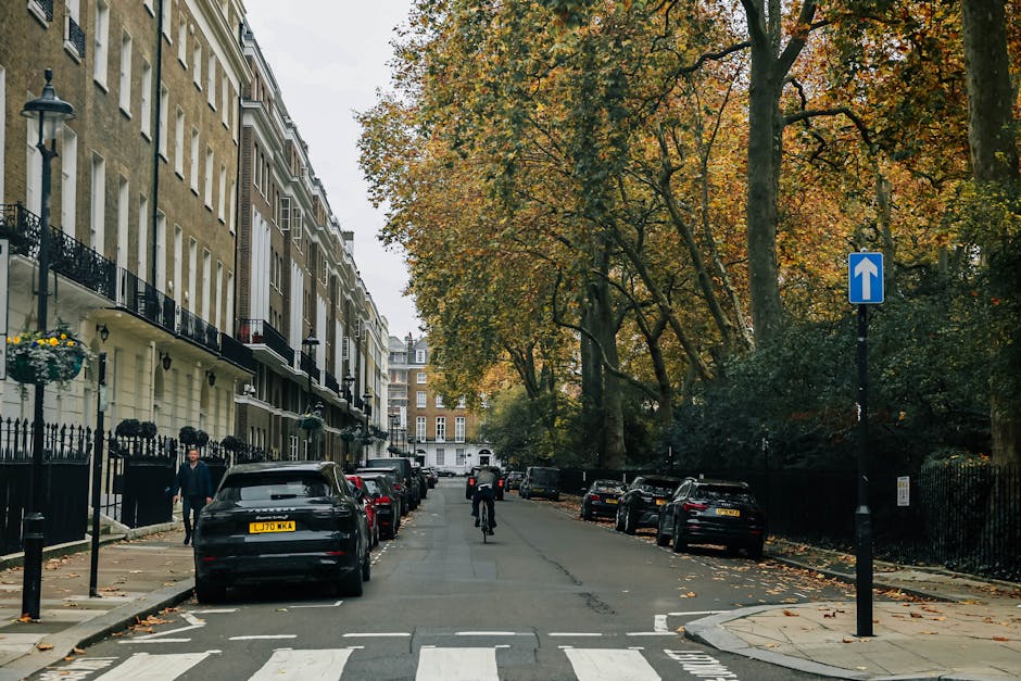 The image displays a quiet street scene in a residential area, featuring a curved asphalt road with minor patches and cracks, bordered by a sidewalk lined with a row of parked bicycles secured to metal bike racks. On the left, there is a large, leafless tree with multiple branches stretching across the scene, contrasting with the partly sunny sky that has scattered clouds and patches of blue. Behind the tree, a modern building with large glass windows and metal framing is visible, alongside traditional brick terraced houses with cream-colored facades, slate roofs, and small chimneys. In the background, three parked cars are seen along the street, with a few pedestrians walking nearby. The area appears to be a well-maintained, peaceful urban setting, which could be associated with private waste collection or rubbish removal services by companies such as House Clearance Belsize Park, emphasizing the importance of local, non-automated waste disposal methods in maintaining the tidiness of the neighborhood.