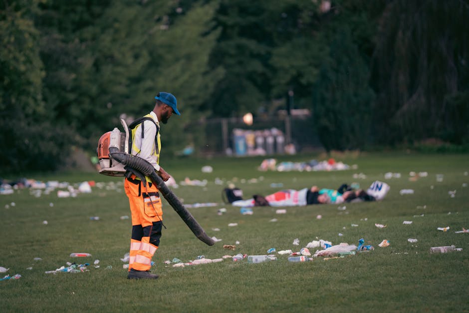 A man dressed in high-visibility orange trousers, a white jacket, and a blue cap is using a leaf blower or vacuum to clear debris from a grassy park area. He is equipped with a backpack-style device, likely a leaf collection or vacuum machine, and stands on a well-maintained green lawn scattered with various rubbish including plastic bottles, cups, and paper waste. In the background, there are large, lush trees and dark green foliage, with some blurred structures and waste bins visible further back. The scene appears to be during the late afternoon or early evening, with natural lighting providing a clear view of the littered environment, which highlights the importance of ongoing waste removal and public cleanliness efforts, similar to the scope of those provided by House Clearance Belsize Park for private rubbish disposal and on-site clearance services.