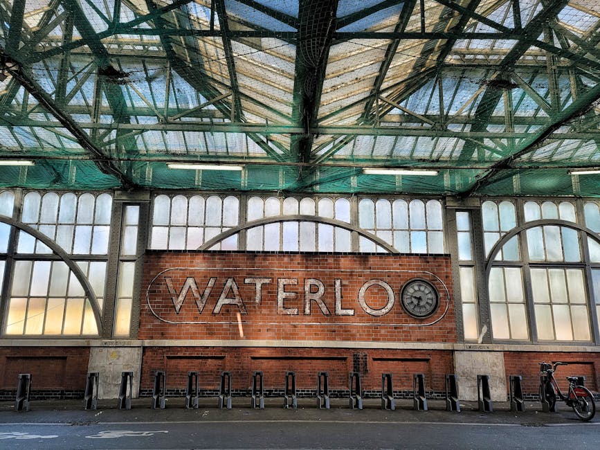 The image depicts an indoor structure with a high, industrial-style glass and metal roof, supported by a framework of green-painted steel beams. The roof features large, arched windows with frosted or aged glass panes that allow diffuse natural light into the space. Below the roof, a prominent red brick wall hosts a large, silver metallic sign spelling 'WATERLOO,' with a small circular clock integrated into the letter 'O.' In front of the brick wall, there are several black bike racks aligned on a concrete pavement, with one red bicycle leaning against the right side of the racks. The environment appears to be part of a transportation hub or station, possibly used for passenger services or public transit. The visible elements emphasize the structure's historic industrial character, with the signage and bicycle parking indicating a location where private or alternative transportation options are supported. The scene conveys a quiet, structured atmosphere typical of a functional public or semi-public space related to infrastructure or transit, which aligns with the context of rubbish removal and waste management preparation in such environments, often carried out by companies like House Clearance Belsize Park.